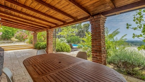 An outdoor patio with a wooden table and chairs, surrounded by greenery and a view of a swimming pool. - Beach front home in Rab (Rab)