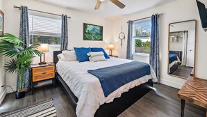 A bedroom with a bed, bedside tables, a ceiling fan, a large window with curtains, a potted plant, and a painting on the wall. - Charming Bungalow w/ Pool in North Palm Beach (West Palm Beach)