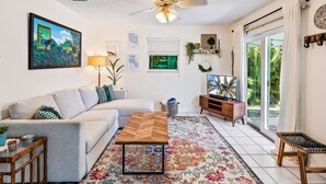 A living room with a sofa, coffee table, TV, and a large window with curtains. - Charming Bungalow w/ Pool in North Palm Beach (West Palm Beach)