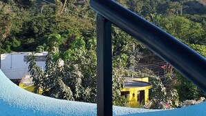 A view from a balcony with a metal railing, overlooking a lush green landscape with a hill and a yellow structure in the background.