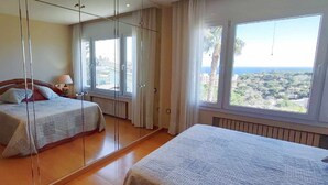 Ein Schlafzimmer mit Bett, Nachttisch mit Lampe, großem Fenster mit Stadtblick und einem Spiegel, der den Raum reflektiert.