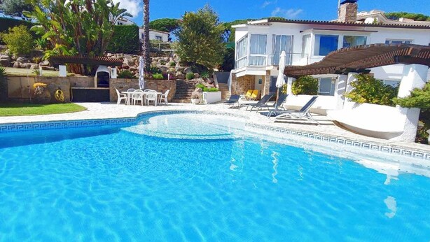 Ein Pool mit gefliestem Rand, umgeben von einem Garten mit verschiedenen Pflanzen und einem Gebäude mit Balkon.