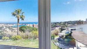 Blick aus einem Fenster auf eine Küstenlandschaft mit einer Palme, einem Swimmingpool und einer Stadt am Horizont.
