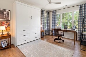 A room with a large white wardrobe, a desk with a chair, a standing lamp, and a window with curtains.