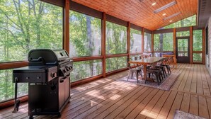 Un espace extérieur abrité, doté d’un barbecue, d’une table à manger, de chaises et de grandes fenêtres offrant une vue sur les arbres.