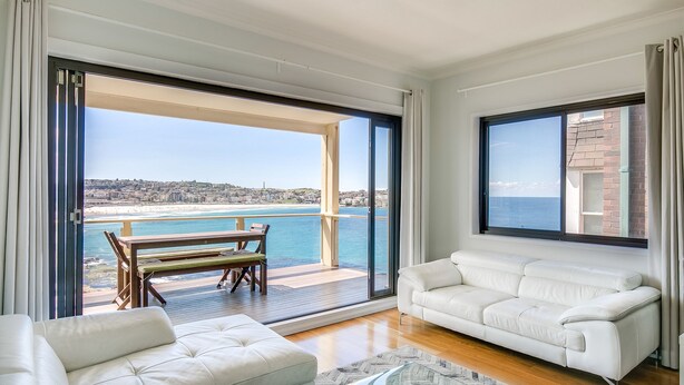 Un salon moderne avec un canapé blanc, une table basse en verre et une terrasse en bois donnant sur une plage.