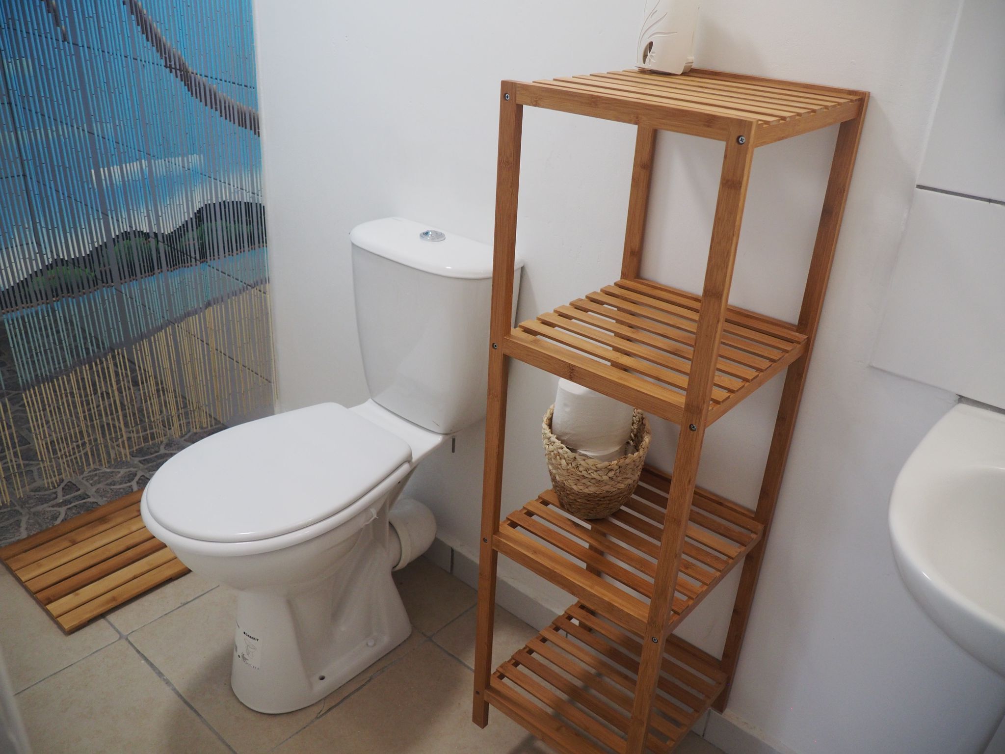 A bathroom with a white toilet, a wooden shelf unit with baskets, and a blue shower curtain.