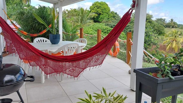 A patio with a red hammock, white chairs, a barbecue grill, and potted plants.