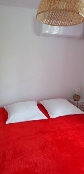 Una cama con dos almohadas blancas y una colcha roja, una mesita de noche blanca con una lámpara y una cesta tejida en la pared.
