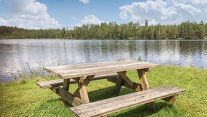 A wooden picnic table with benches by a calm lake, surrounded by lush greenery and trees.