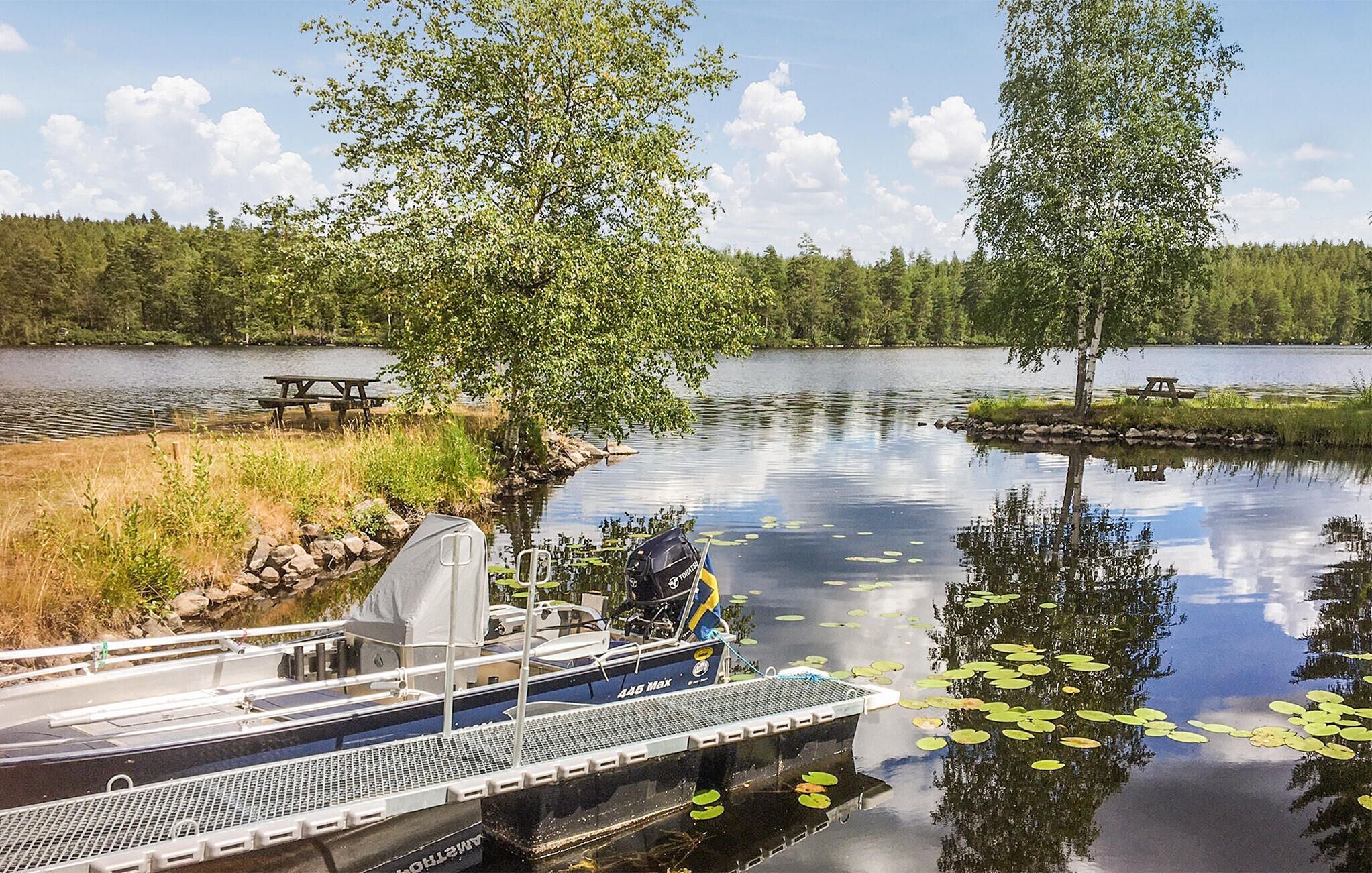 Ein Boot liegt an einem ruhigen See, im Hintergrund sind Picknicktische und Bäume zu sehen.