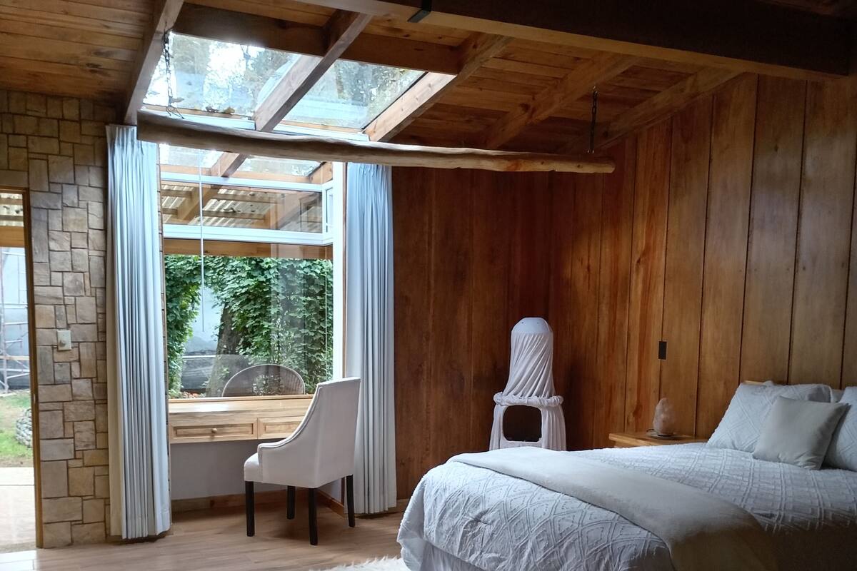 A bedroom with a bed, a chair, a window, and a view of greenery outside.