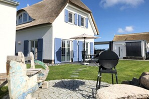 A house with a thatched roof, blue shutters, and a barbecue grill in the yard. - Villa Morgensünn - House in an idyllic location with sauna, terrace and garden - (Trent)