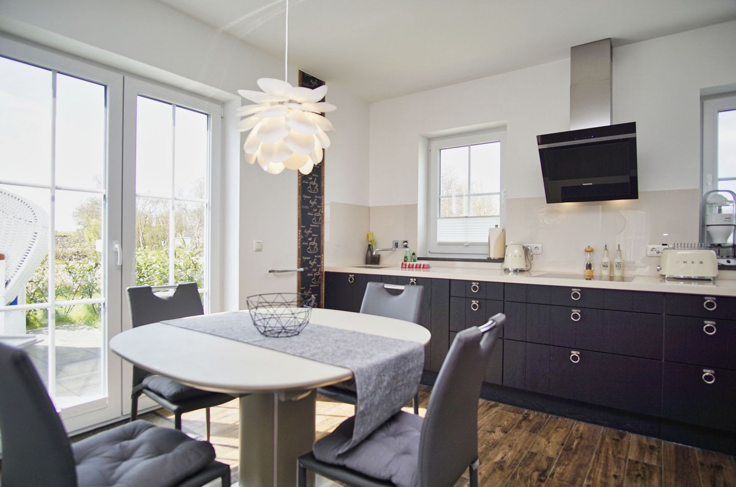 A modern kitchen with dark cabinets, a white countertop, and a built-in oven. There is a dining area with a round table and chairs, and a large window providing natural light.