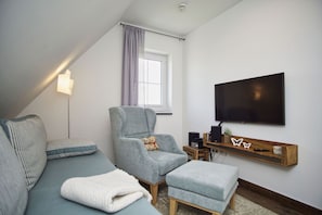 A modern living room with a grey sofa, a grey armchair, a white ottoman, a flat-screen TV mounted on the wall, and a small wooden side table with speakers and a plant.