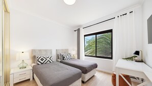 Une chambre moderne avec deux lits, un bureau blanc et une grande fenêtre donnant sur des palmiers.