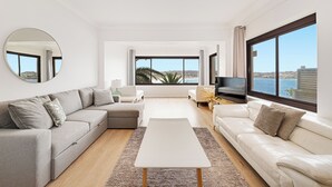 Un salon moderne avec un canapé d’angle, une table basse blanche et un téléviseur. De grandes fenêtres offrent une vue sur la mer et le paysage urbain.