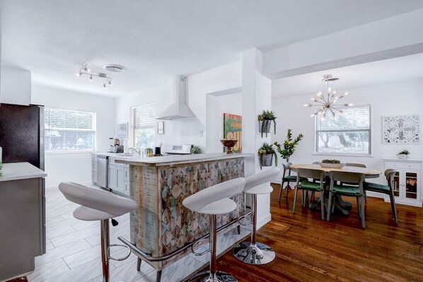 A modern kitchen with a central island, bar stools, a dining area with a table and chairs, and a chandelier.