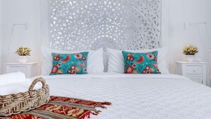 Une chambre à coucher avec un lit blanc, des coussins à motifs floraux, un panier en osier et une tête de lit décorative.