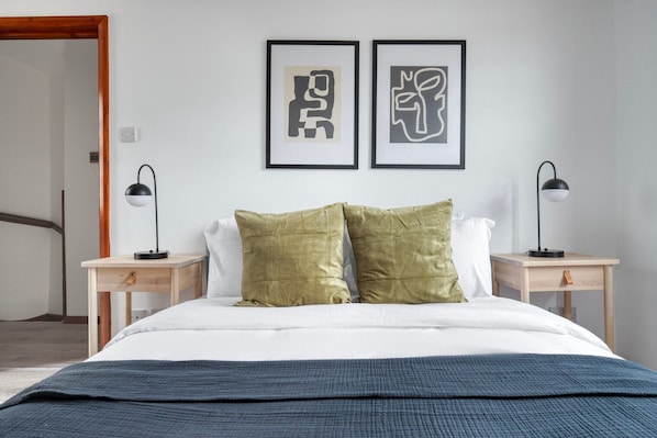 A bedroom with a bed, two bedside tables, two lamps, and two abstract art pieces on the wall.