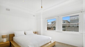 Un dormitorio con cama de madera, ropa de cama blanca, una mesita de noche con lámpara y ventana con vista a edificios.