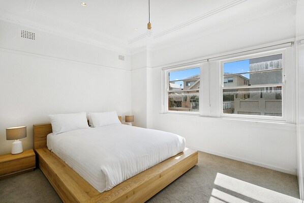 A bedroom with a wooden bed, white bedding, a nightstand with a lamp, and a window with a view of buildings.