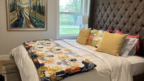 Ein Schlafzimmer mit einem großen Bett, einem gepolsterten Kopfteil, einem Fenster mit Blick ins Grüne und einem Bild an der Wand.