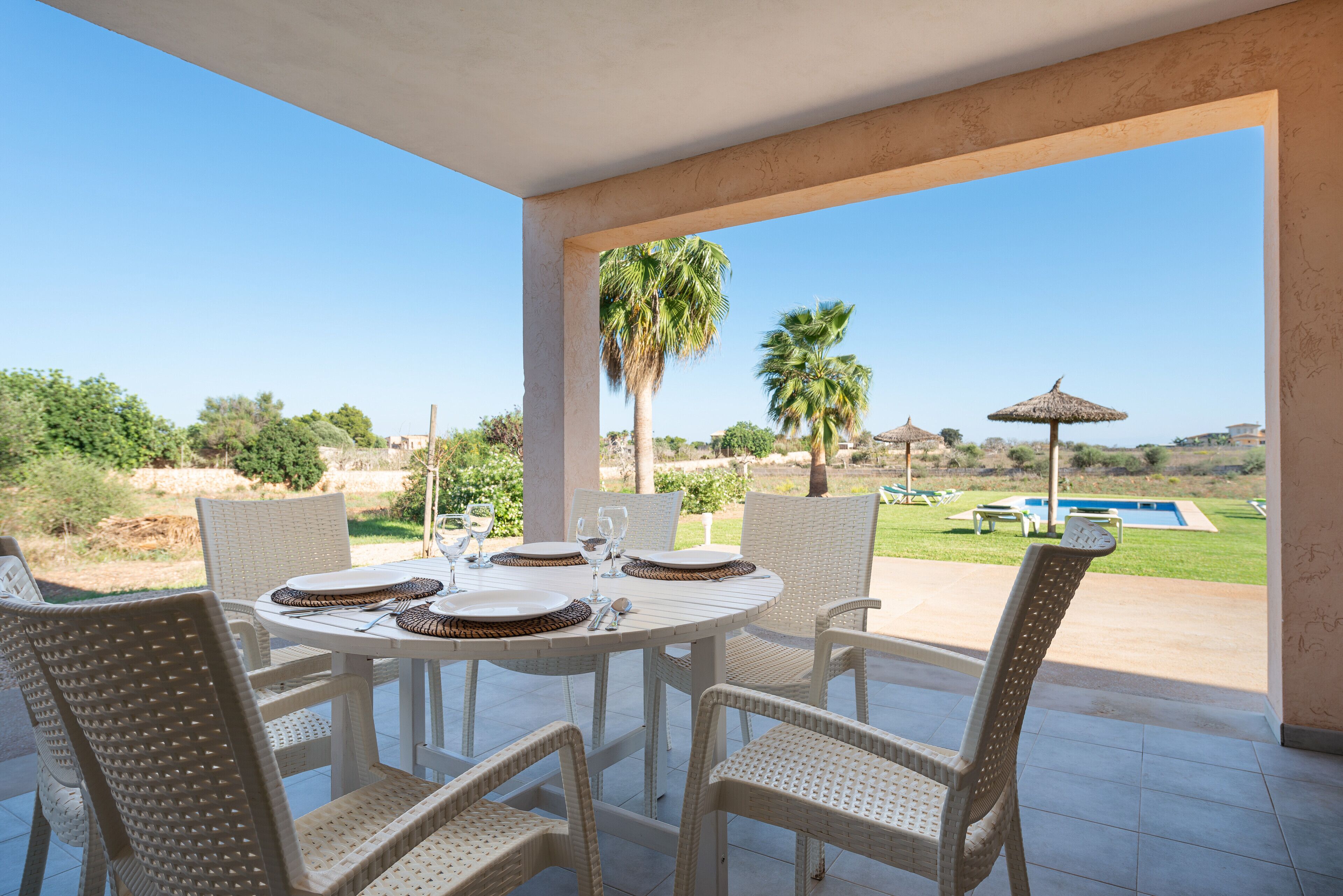 Un área de comedor al aire libre techada con una mesa dispuesta para cuatro personas, rodeada de sillas de mimbre, con vista a una piscina y palmeras.