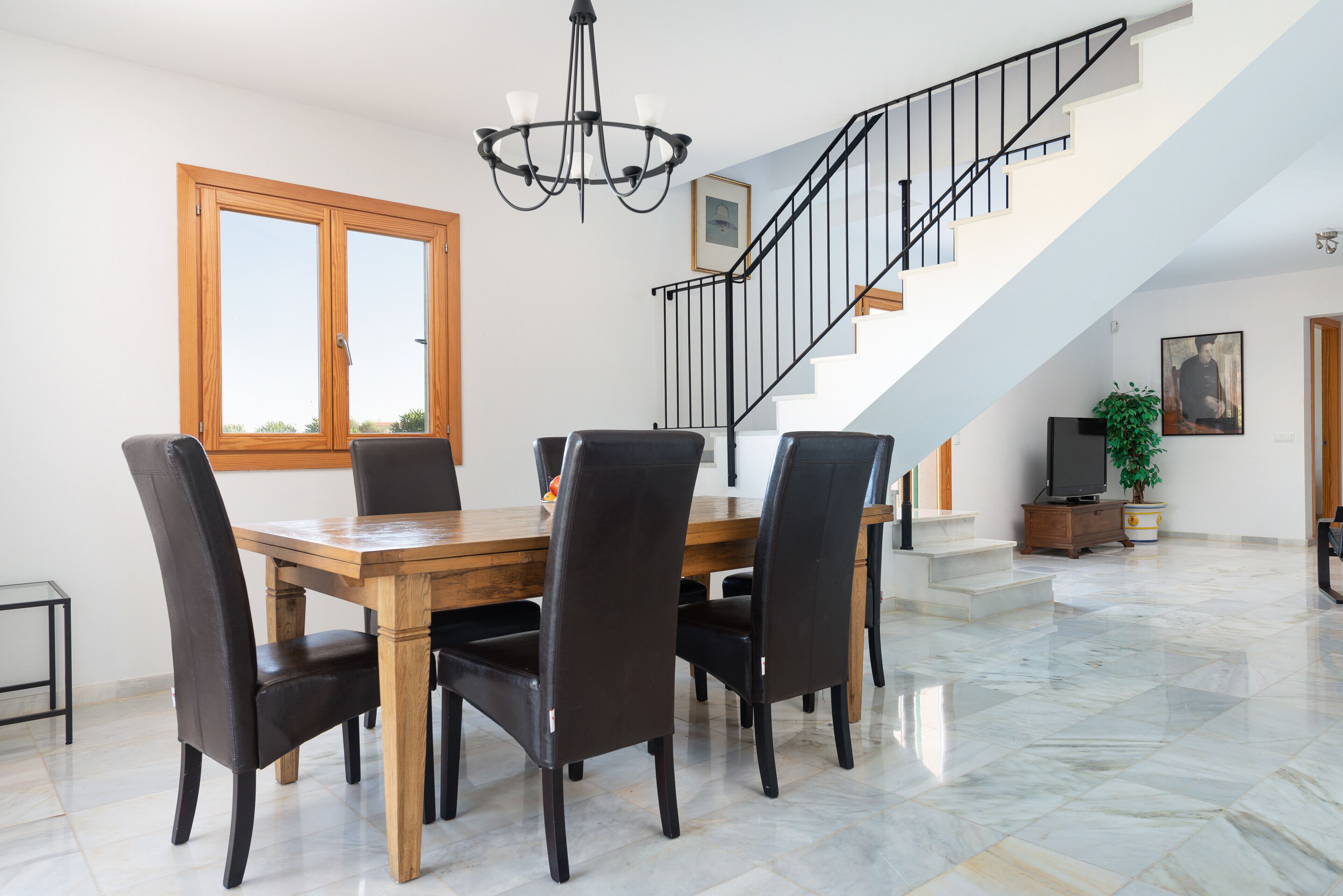 Un comedor con una mesa y sillas de madera, una araña y una escalera con barandilla.