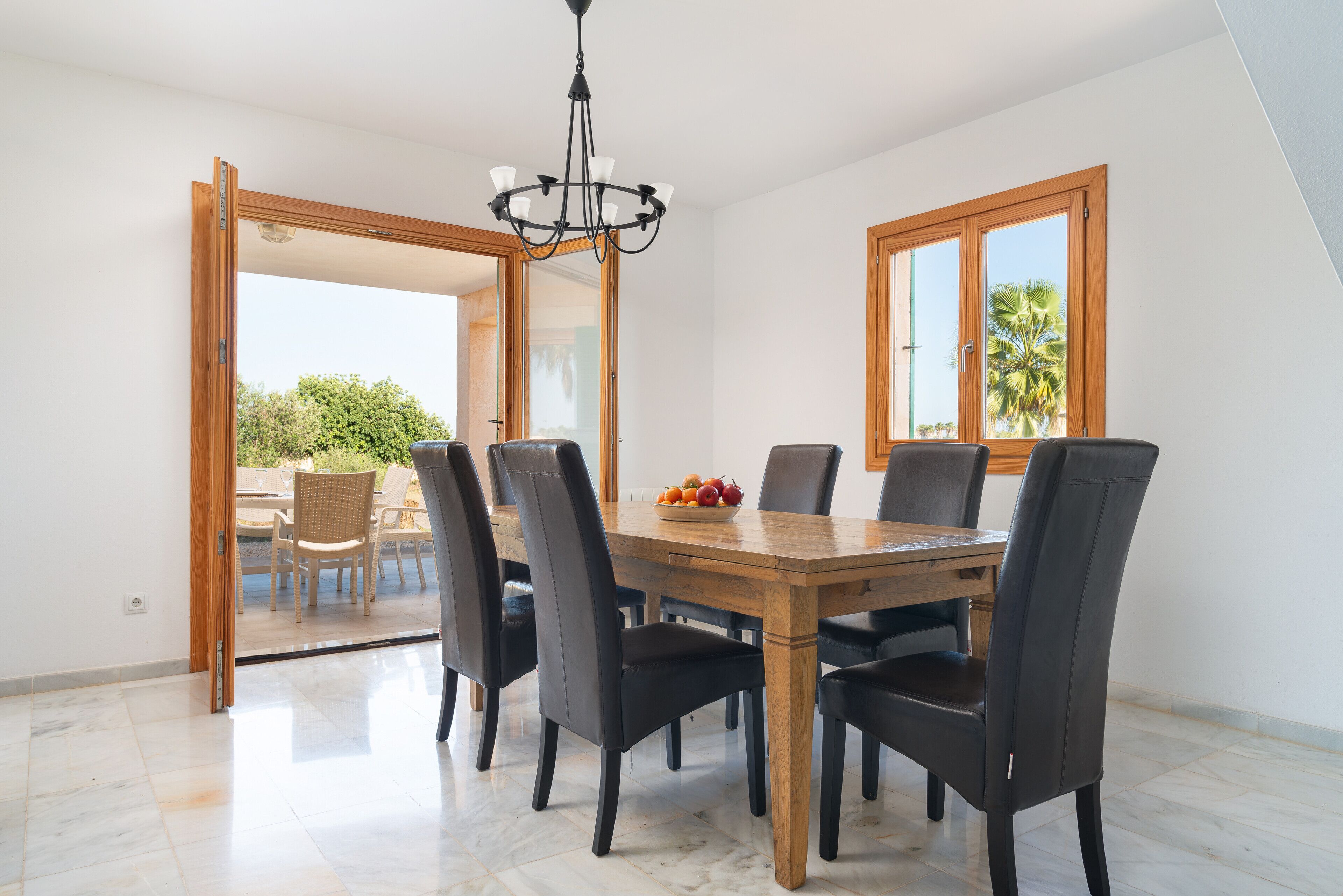 Un comedor con una mesa de madera y seis sillas, una lámpara de araña y vistas a un patio exterior a través de un ventanal abierto.