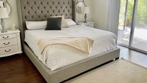 A bedroom with a tufted headboard, a large bed with white bedding, two bedside tables with lamps, and a view of greenery through a sliding glass door.
