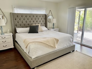 Quarto com cabeceira estofada, cama grande com roupa de cama branca, duas mesas de cabeceira com abajures e vista para o verde através de uma porta de vidro deslizante.