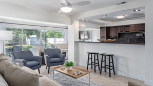 A modern kitchen with dark cabinets, a microwave, and a countertop. There is a ceiling fan, a bar stool, and a window overlooking a garden.