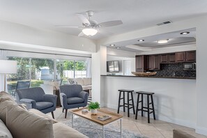 Cozinha moderna com armários escuros, micro-ondas e bancada. Há um ventilador de teto, um banquinho de bar e uma janela com vista para um jardim.