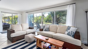 A spacious living room with a large sofa, a coffee table, and a wicker chair. There are large windows with curtains, and a view of the outdoors.