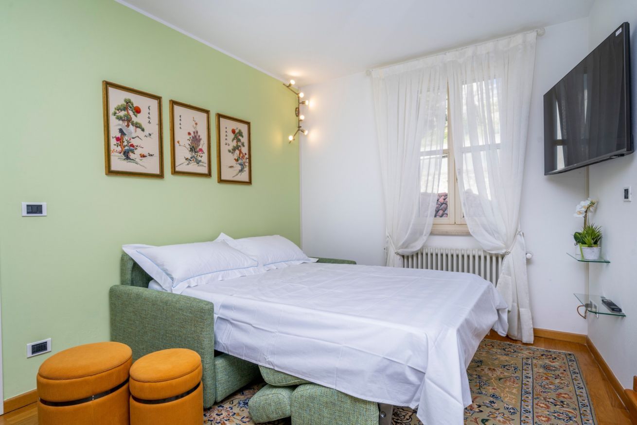 A bedroom with a bed, green upholstered armchair, two ottomans, a radiator, a TV, and artwork on the wall.