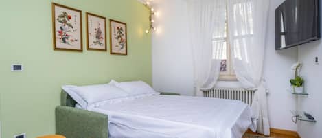 A bedroom with a bed, green upholstered armchair, two ottomans, a radiator, a TV, and artwork on the wall.