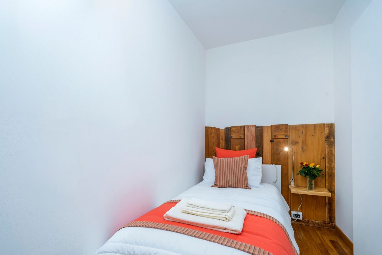 A bedroom with a wooden headboard, a bed with white and red bedding, a bedside table with a lamp, and a small plant.