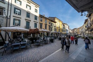 Une rue pavée avec des tables et des chaises installées à l’extérieur, des bâtiments avec des volets et un ciel dégagé.