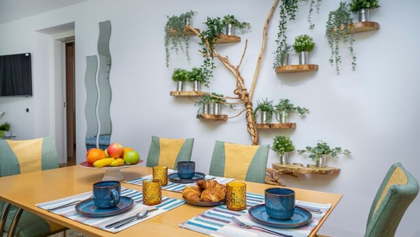 A dining area with a wooden table, chairs, and a breakfast setting including fruits, croissants, and blue cups. - GuestHost - Mercatovecchio Ivy House (Udine)