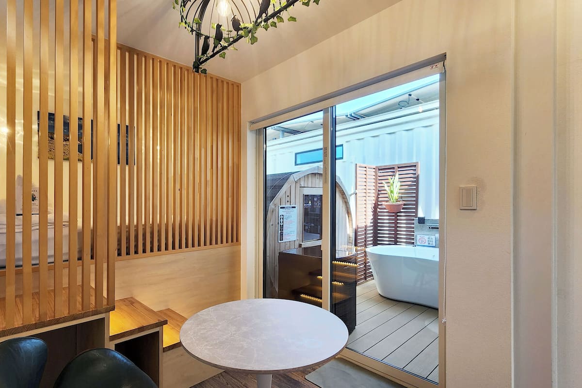 A room with a wooden partition, a round table, and a view through a glass door to an outdoor area with a white structure.