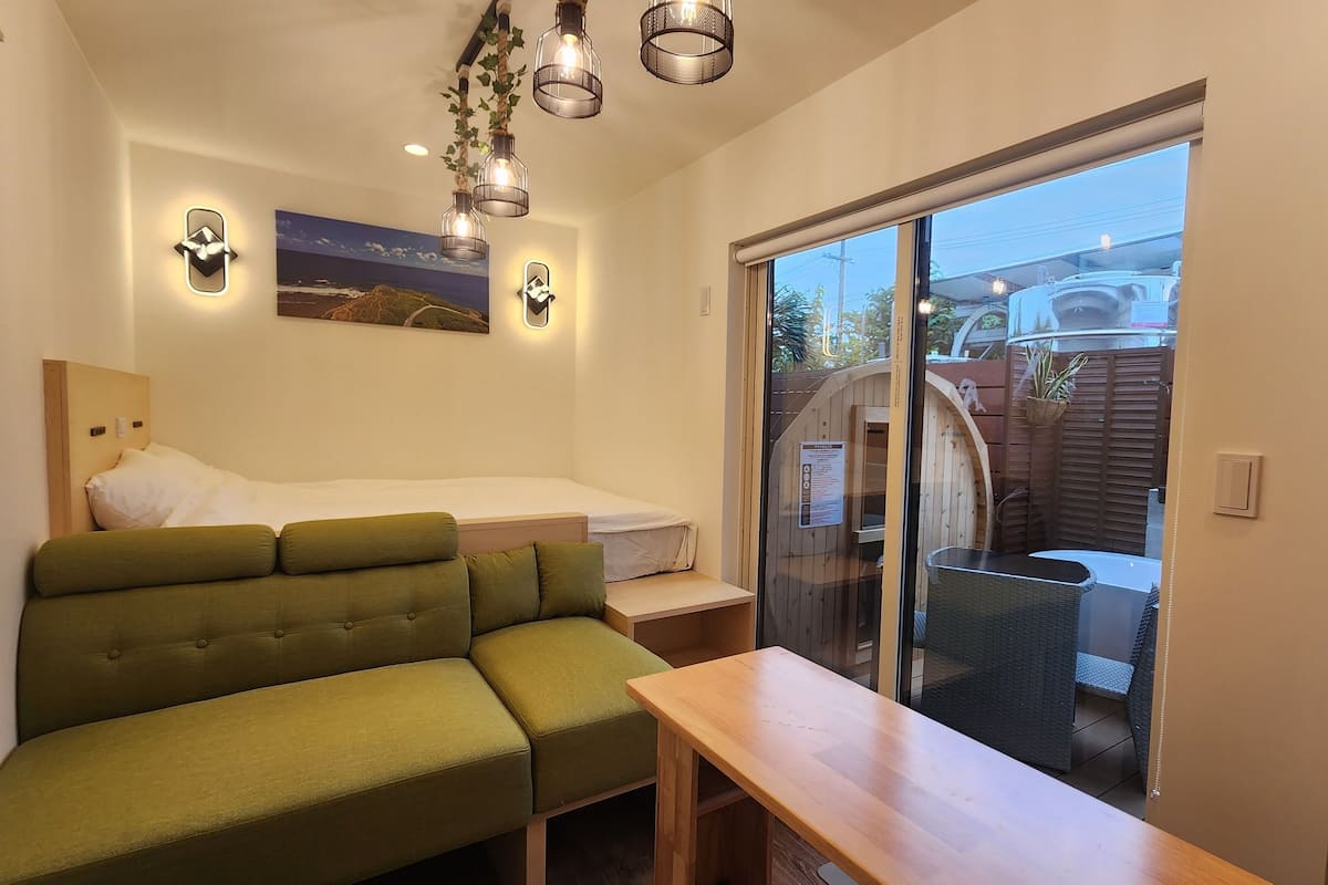 A compact room with a green sofa, a small bed, a wooden table, and a sliding glass door leading to an outdoor area.