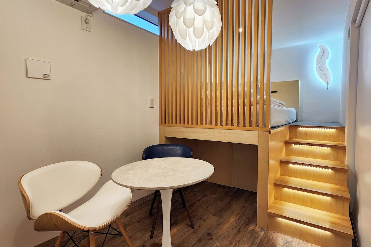 A small, cozy room with a wooden desk, a round table, two chairs, a slanted ceiling with a light fixture, and a staircase with illuminated steps.