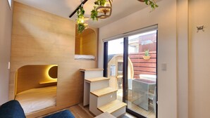 A compact room with a built-in bed, wooden shelves, a small staircase, and a sliding glass door leading to an outdoor area with a wicker chair and a table. - ZUMI TERRACE UCHINAAMIYA RESORT (Miyakojima)
