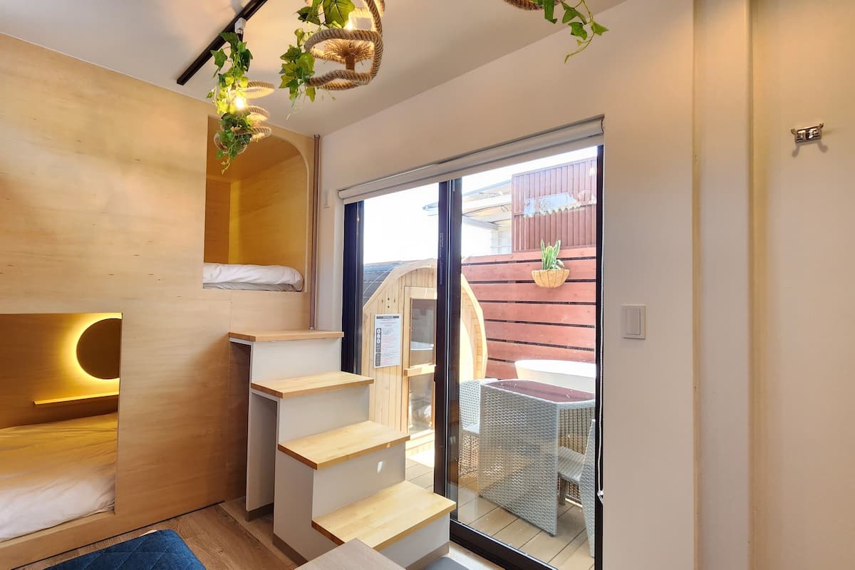 A compact room with a built-in bed, wooden shelves, a small staircase, and a sliding glass door leading to an outdoor area with a wicker chair and a table.
