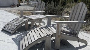 Adirondack-stoelen en een kleine tafel op een betonnen terras, met een grindrand en planten op de achtergrond.