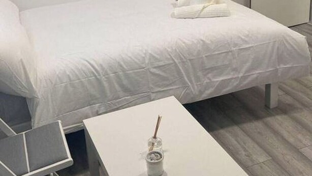 A modern hotel room with a neatly made bed, a small white table with a drink, and a chair.