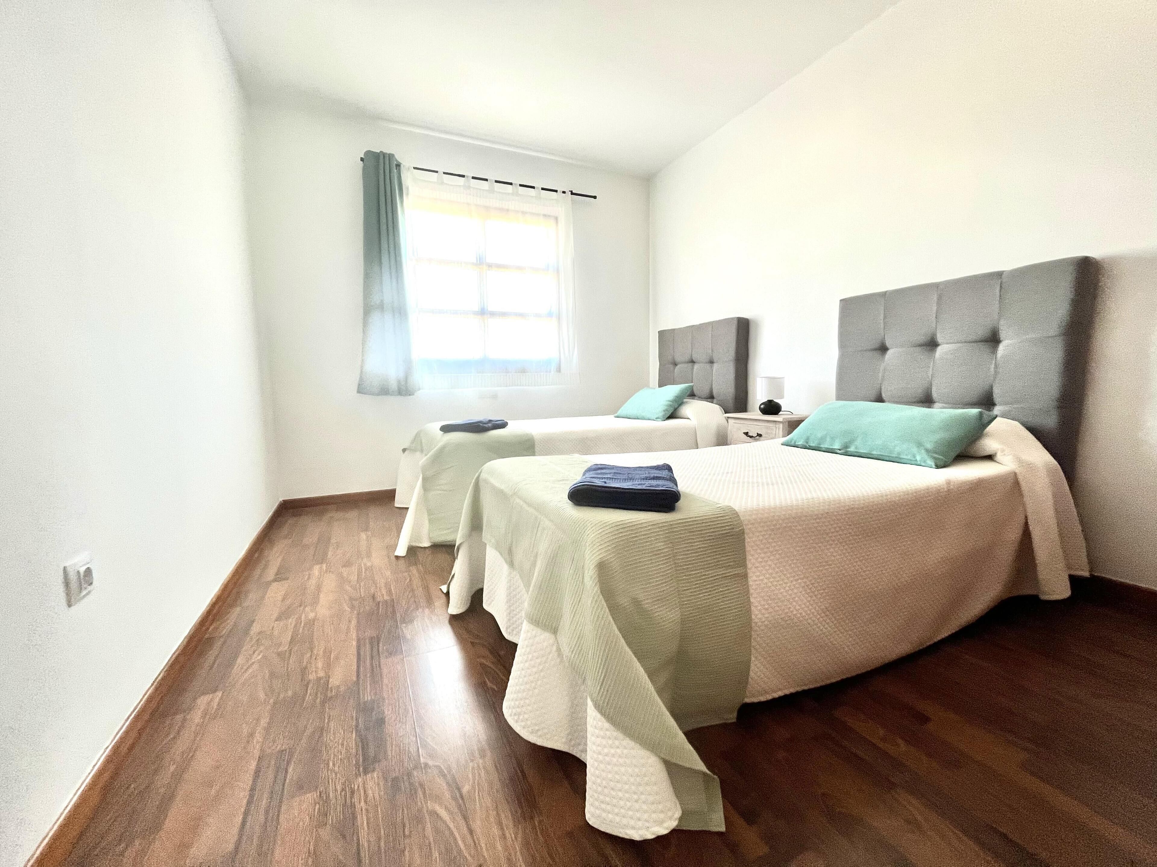 Een hotelkamer met twee bedden, houten vloeren, een raam met gordijnen en een nachtkastje met een lamp.