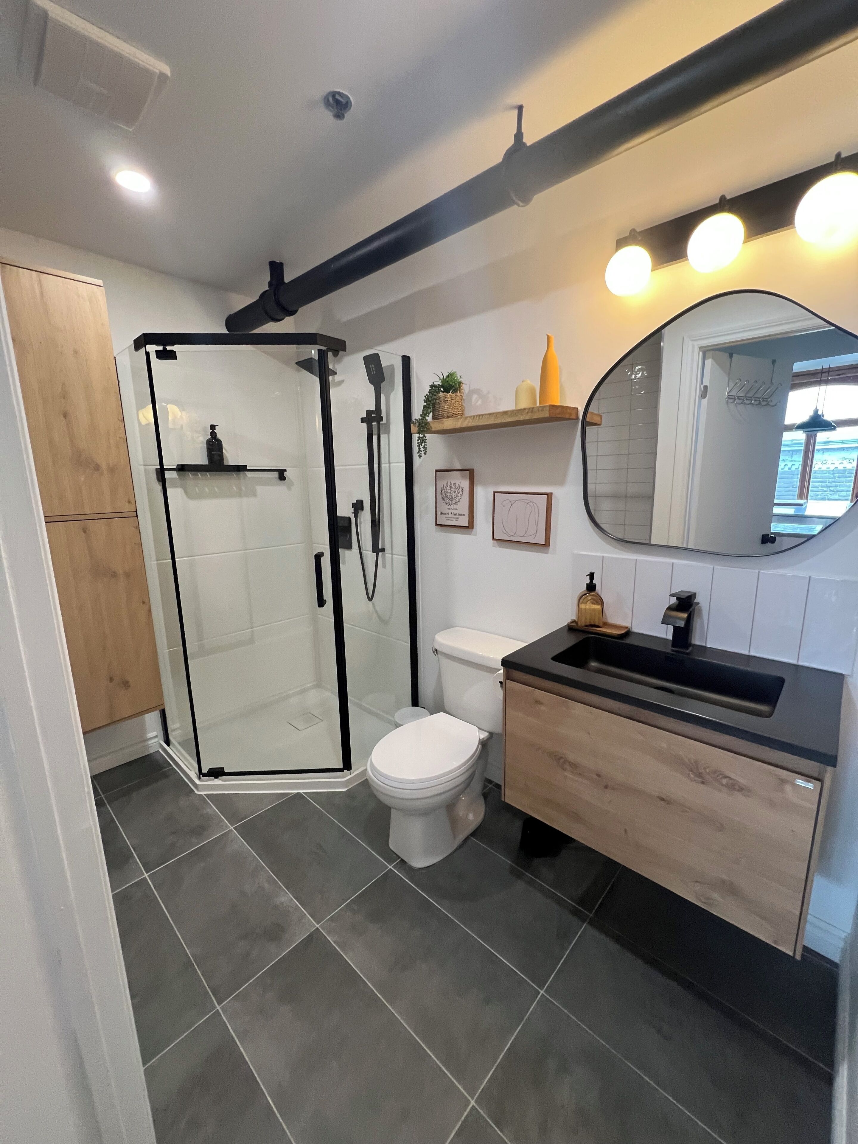 Une salle de bain moderne avec une douche à parois de verre, des toilettes blanches et un meuble-lavabo en bois doté d’un plan de travail noir.