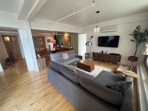 A modern living room with a grey sofa, wooden flooring, a flat-screen TV, and a dining area with a table and chairs.